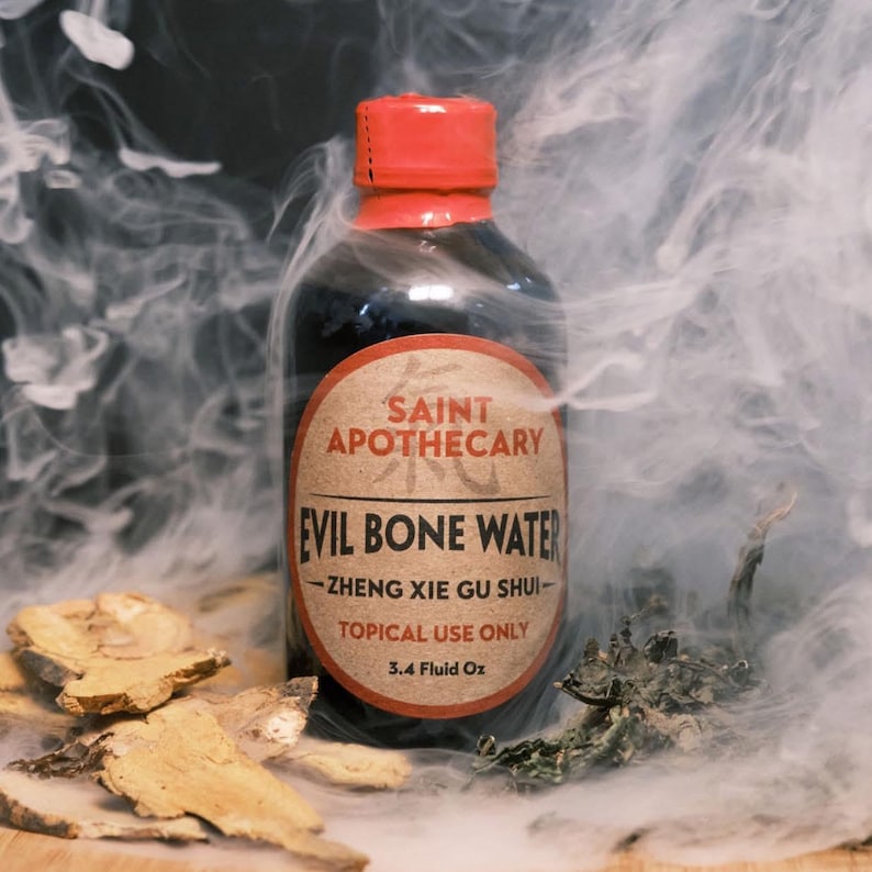 May include: A dark glass bottle with a red cap, labeled "SAINT APOTHECARY EVIL BONE WATER." The label includes "ZHENG XIE GU SHUI" and "TOPICAL USE ONLY, 3.4 Fluid Oz." The image has a smoky background with herbs.