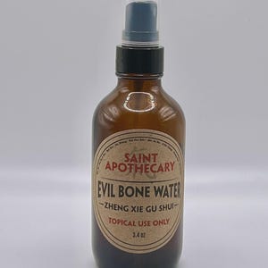 May include: A brown glass bottle with a black spray nozzle. The label reads "Saint Apothecary Evil Bone Water - Zheng Xie Gu Shui - Topical Use Only" and contains 3.4 oz of liquid. The bottle is against a white background.