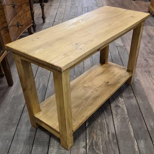 Könnte beinhalten: Ein hellfarbiger, rechteckiger Holztisch mit einem unteren Regal. Der Tisch hat ein schlichtes, rustikales Design mit sichtbarer Holzmaserung und einer natürlichen Oberfläche. Der Tisch ist etwa 122 cm lang.