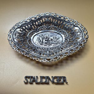 Könnte beinhalten: Eine ovale Schale aus silberfarbenem Metall mit einem filigranen Gitterrand und Blumenmotiven. In der Mitte der Schale befindet sich ein Relief mit drei Putten. Das Wort STAUDINGER steht in Metallbuchstaben unter der Schale.