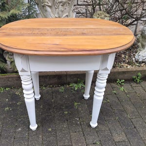 Könnte beinhalten: Ovaler Holztisch mit hellbrauner Tischplatte und weißem Untergestell. Der Tisch hat vier gedrechselte Beine und eine kleine Schublade. Die Tischplatte hat einen Durchmesser von ca. 76 cm.