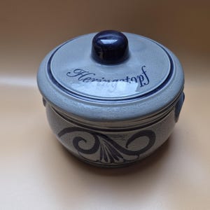 Könnte beinhalten: Ein runder, grauer Keramiktopf mit Deckel und dunkelblauem Knopf. Der Deckel trägt den Schriftzug "Herringstopf" in blauer Schrift. Der Topf hat ein dekoratives blaues Wirbeldesign um den Boden.