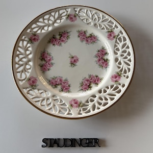 Könnte beinhalten: Ein weißer Porzellanteller mit goldenem Rand und filigranem Lochmuster. Der Teller zeigt Gruppen von rosa Rosen. Das Wort "STAUDINGER" befindet sich unter dem Teller.