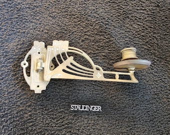 May include: A gold-toned metal wall sconce with an ornate design. The sconce has a single light fixture and a decorative element that resembles a stylized wave. The sconce is labeled "STAUDINGER".