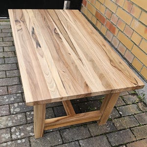 Könnte beinhalten: Ein Holz-Couchtisch mit natürlicher Holzoptik und einem einfachen, rechteckigen Design. Der Tisch hat eine breite, flache Tischplatte und ein stabiles Gestell mit einer kreuzförmigen Stütze.