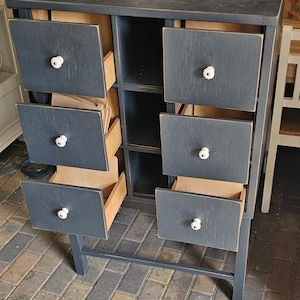 Könnte beinhalten: Ein dunkelgrauer Holzschrank mit sechs Schubladen und einem Regal. Die Schubladen haben weiße Knöpfe und sind teilweise geöffnet, wodurch das natürliche Holzinterieur sichtbar wird. Der Schrank wird von Beinen getragen und steht auf einem Ziegelboden.