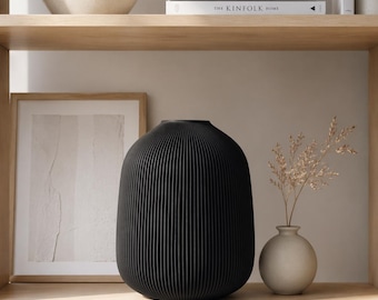 May include: A black, ribbed vase with a rounded shape sits on a wooden shelf, alongside a small, beige vase holding dried stems. A framed artwork and books titled "KINFOLK TRAVEL" and "THE KINFOLK HOME" are also visible.