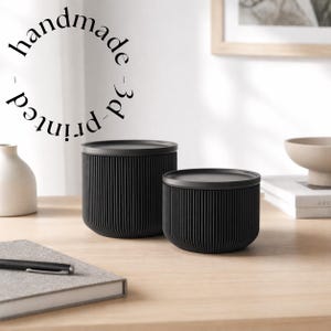 Könnte beinhalten: Zwei schwarze, handgefertigte 3D-gedruckte Behälter mit Deckeln. Die Behälter haben eine gerippte Textur und sind zylindrisch. Der Text "handmade - 3d printed" ist kreisförmig angeordnet.