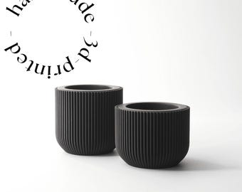Puede incluir: Dos macetas negras impresas en 3D con detalles de costillas verticales. Las macetas son cilíndricas con fondos redondeados. El texto "handmade 3d printed" está dispuesto en círculo encima de las macetas.