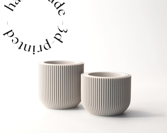 Puede incluir: Dos macetas cilíndricas acanaladas de color gris claro, impresas en 3D. Las macetas tienen una base redondeada y son de diferentes tamaños. El texto "handmade 3d printed" está en un círculo encima de las macetas.