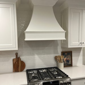 May include: White kitchen range hood with a curved design, installed above a stainless steel stove with a black cooktop and oven. The stove has a digital display showing "72."