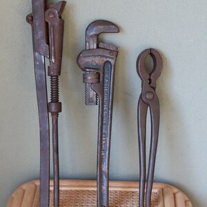 Może przedstawiać: Trzy zabytkowe narzędzia metalowe: klucz do rur, klucz do rur z napisem "THE RIDGE TOOL CO. ELYRIA OHIO U.S.A. 18" i szczypce. Narzędzia są ułożone pionowo na powierzchni z plecionego wikliny.