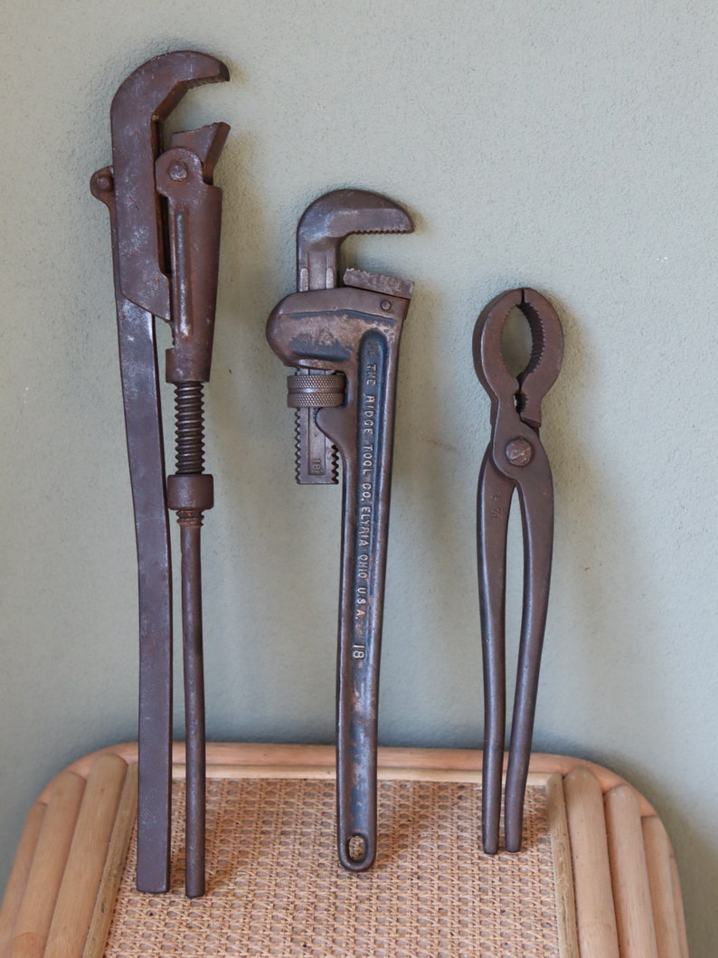 Może przedstawiać: Trzy zabytkowe narzędzia metalowe: klucz do rur, klucz do rur z napisem "THE RIDGE TOOL CO. ELYRIA OHIO U.S.A. 18" i szczypce. Narzędzia są ułożone pionowo na powierzchni z plecionego wikliny.