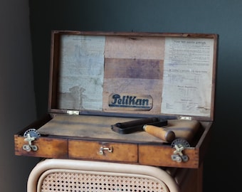 May include: An antique wooden box with a hinged lid, containing a roller and a metal tool. The box is labeled "Pelikan" and has instructions for use in Dutch.