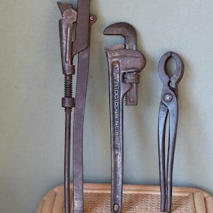 Może przedstawiać: Trzy stare, zardzewiałe metalowe klucze, dwa to klucze do rur, a jeden to szczypce. Klucze do rur mają napis "TRADE RIDGID MARK PAT. 1727223 18".
