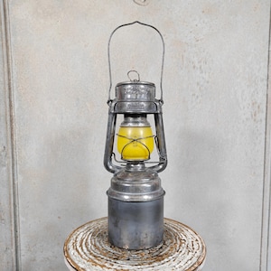 Può includere: Una lanterna in metallo vintage con un globo di vetro giallo e un manico. La lanterna ha una finitura argentata ed è appoggiata su una superficie di legno. Il design ricorda l'illuminazione tradizionale per il campeggio o le attività all'aperto.