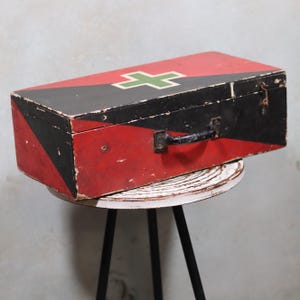 Op de afbeelding: Een vintage houten EHBO-kist geschilderd in rood, zwart en wit. De bovenkant heeft een groen kruis op een rode achtergrond. De doos heeft een zwarte handgreep en een metalen sluiting. Hij staat op een kleine, ronde, witte tafel.