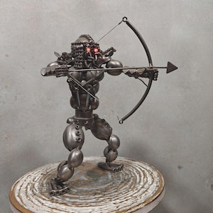 Puede incluir: Una escultura de metal de una figura sosteniendo un arco y una flecha. La figura está hecha de varias piezas de metal, incluyendo tuercas, tornillos y arandelas. La escultura está pintada en un color plateado metálico.