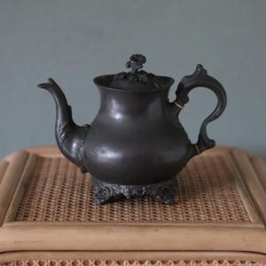 以下が含まれることがあります： 蓋に花柄の装飾が施された黒い錫製のティーポット。湾曲した取っ手と、編み込まれた表面の上に置かれています。