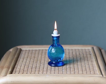 May include: A blue glass oil lamp with a white wick and a flame burning. The lamp is sitting on a woven wicker surface.