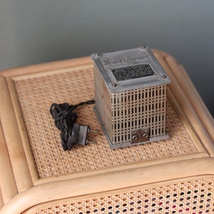 Könnte beinhalten: Ein silberner Metalltransformator mit einem schwarzen Kabel und Stecker. Der Transformator ist mit "Transformator" beschriftet und hat ein Gittermuster auf der Vorderseite.