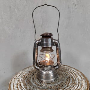 Op de afbeelding: Een zilverkleurige metalen stormlantaarn met een heldere glazen bol die een kleine, brandende vlam beschermt. De lantaarn heeft een draadhandvat aan de bovenkant, een ronde voet en een metalen frame met zichtbare structurele onderdelen.