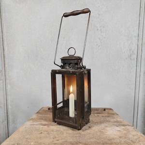 May include: A rectangular metal lantern with a dark, weathered finish, clear glass panels, and a tall wire handle featuring a dark wooden grip. Inside, a single white taper candle stands upright, emitting a small, warm, glowing flame.