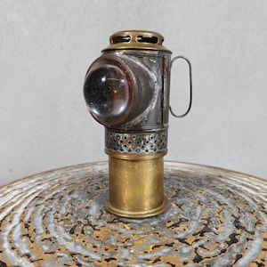 Op de afbeelding: Een antieke lantaarn met een heldere, afgeronde lens en een messing voet. De lantaarn heeft een handvat en een geperforeerd gedeelte nabij de bovenkant. Het metaal heeft een verweerde uitstraling, wat duidt op leeftijd en gebruik. Het ontwerp doet denken aan vintage spoorweg- of maritieme verlichting.