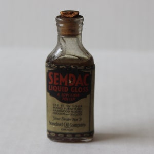 May include: A small glass bottle with a cork stopper and a label that reads "SEMDAC LIQUID GLOSS A SUPERIOR POLISH". The label also includes instructions for use and the company name "Standard Oil Company, Chicago".