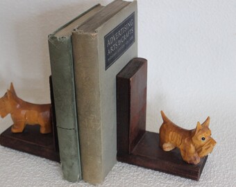 May include: A pair of wooden bookends shaped like Scottish Terriers. The bookends are brown and have a natural wood finish. The bookends are holding up two books, one of which is titled "Advertising Arts & Crafts".