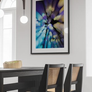 May include: A black framed abstract art print with a blue, purple and yellow colour scheme. The print is hanging on a white wall above a black dining table with two black chairs.