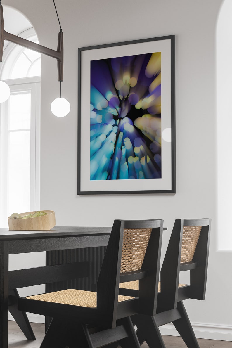 May include: A black framed abstract art print with a blue, purple and yellow colour scheme. The print is hanging on a white wall above a black dining table with two black chairs.