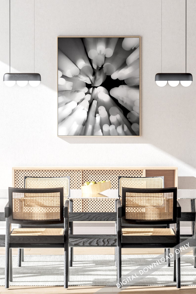 May include: A black and white abstract photograph of a circular pattern of light and dark shapes. The photograph is framed in a thin, wooden frame and is hanging on a white wall above a dining table with two chairs.