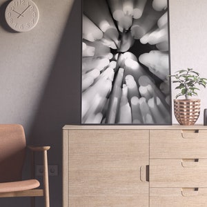 May include: A black and white photograph of a blurred abstract design with a circular pattern of light and dark shapes. The image is framed in a black frame and is hanging on a wall above a wooden cabinet.