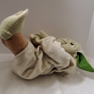 Op de afbeelding: Een pluche speelgoed van Yoda uit Star Wars, liggend op zijn rug met zijn armen gekruist. Het speelgoed draagt een witte robe en heeft groene huid, grote oren en een lange, puntige neus.