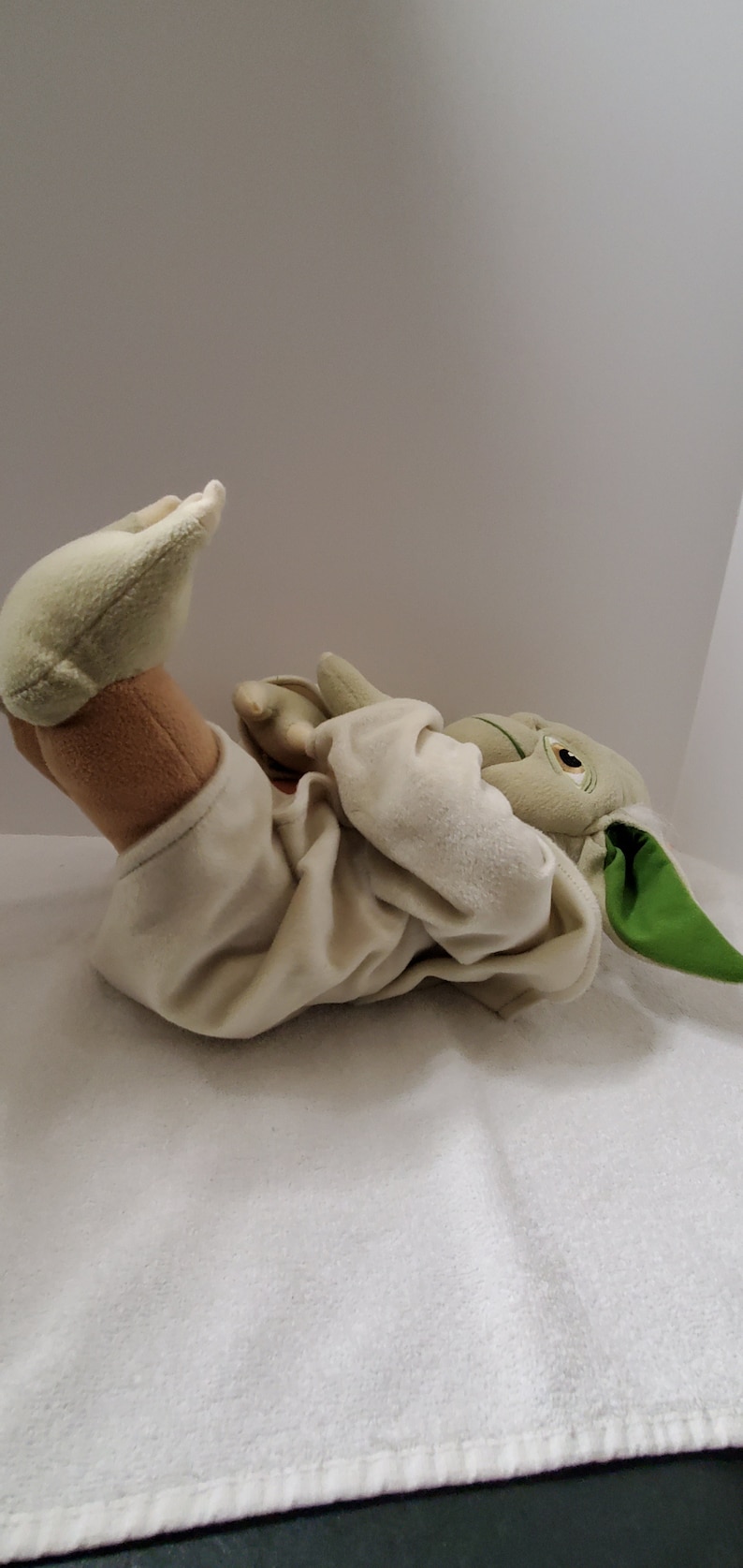 Op de afbeelding: Een pluche speelgoed van Yoda uit Star Wars, liggend op zijn rug met zijn armen gekruist. Het speelgoed draagt een witte robe en heeft groene huid, grote oren en een lange, puntige neus.