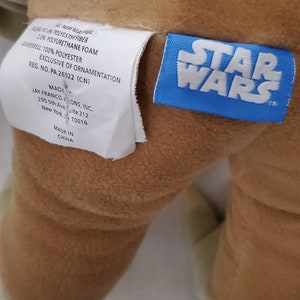 Op de afbeelding: Een bruin pluche speelgoed met een wit label waarop staat "ALL NEW MATERIAL 97.4% POLYESTER FIBER 1.6% POLYURETHANE FOAM COVERSHELL: 100% POLYESTER EXCLUSIVE OF ORNAMENTATION REG. NO. PA 26922 (CN) MADE BY: JAY FRANCO & SONS, INC. 295 5th Ave Suite 312 New York, 10016 MADE IN CHINA" en een blauw label met de tekst "STAR WARS".