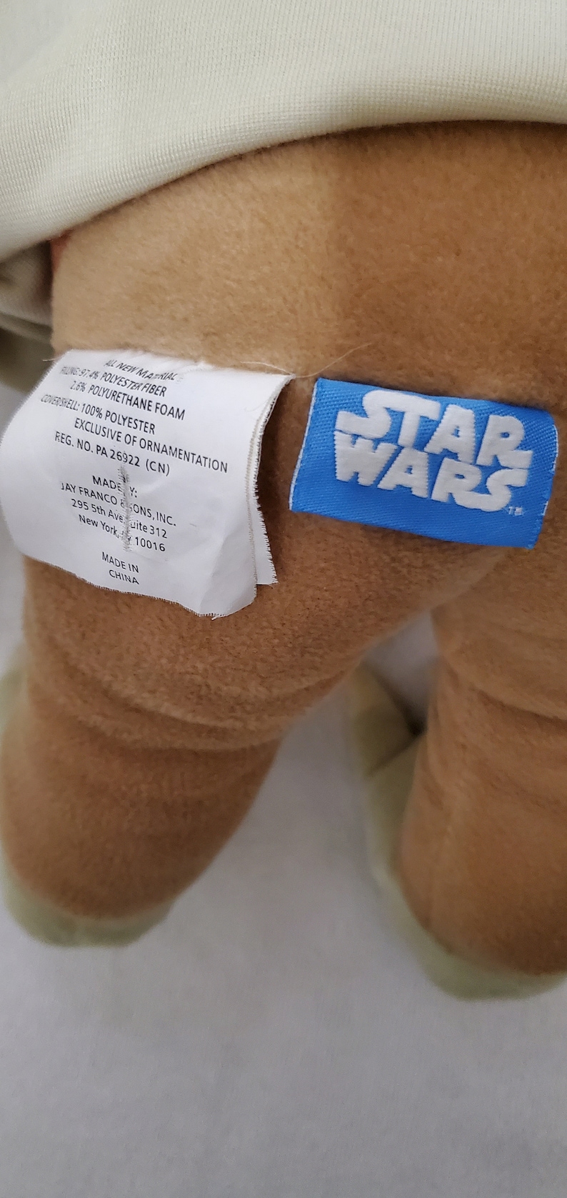 Op de afbeelding: Een bruin pluche speelgoed met een wit label waarop staat "ALL NEW MATERIAL 97.4% POLYESTER FIBER 1.6% POLYURETHANE FOAM COVERSHELL: 100% POLYESTER EXCLUSIVE OF ORNAMENTATION REG. NO. PA 26922 (CN) MADE BY: JAY FRANCO & SONS, INC. 295 5th Ave Suite 312 New York, 10016 MADE IN CHINA" en een blauw label met de tekst "STAR WARS".