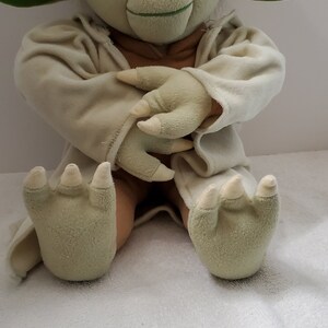 Op de afbeelding: Een groen pluche Yoda-speelgoed dat een witte robe draagt, zittend met gekruiste armen en een glimlach op zijn gezicht.