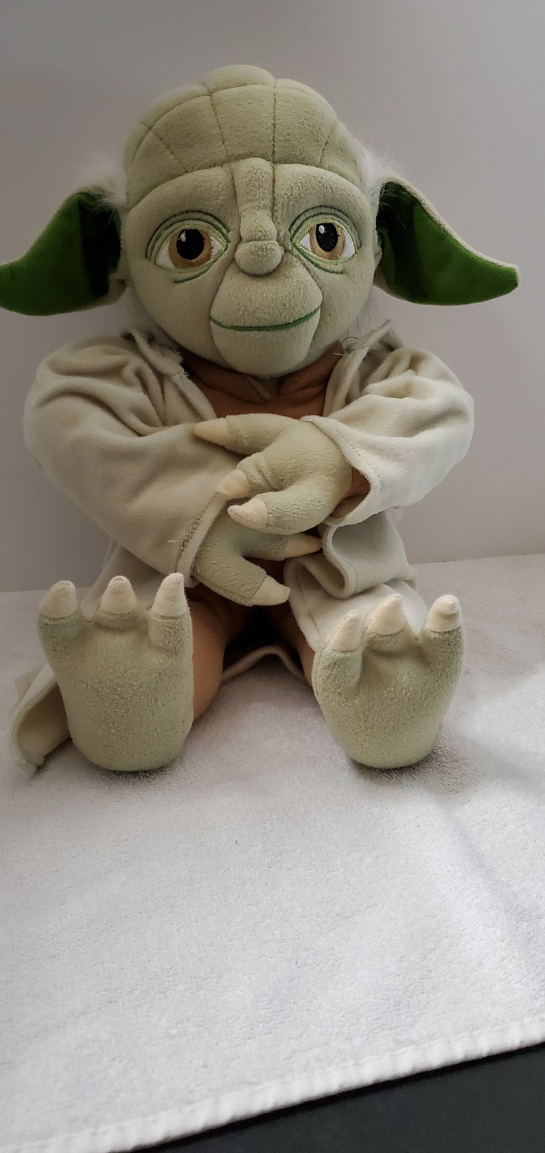 Op de afbeelding: Een groen pluche Yoda-speelgoed dat een witte robe draagt, zittend met gekruiste armen en een glimlach op zijn gezicht.