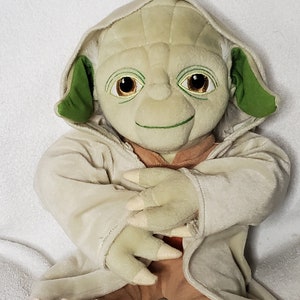 Op de afbeelding: Een groen en bruin pluche speelgoed van Yoda uit Star Wars. Het speelgoed draagt een beige gewaad en heeft een grote glimlach.