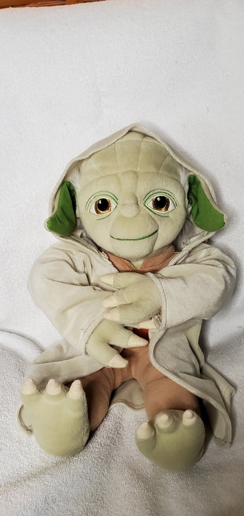 Op de afbeelding: Een groen en bruin pluche speelgoed van Yoda uit Star Wars. Het speelgoed draagt een beige gewaad en heeft een grote glimlach.