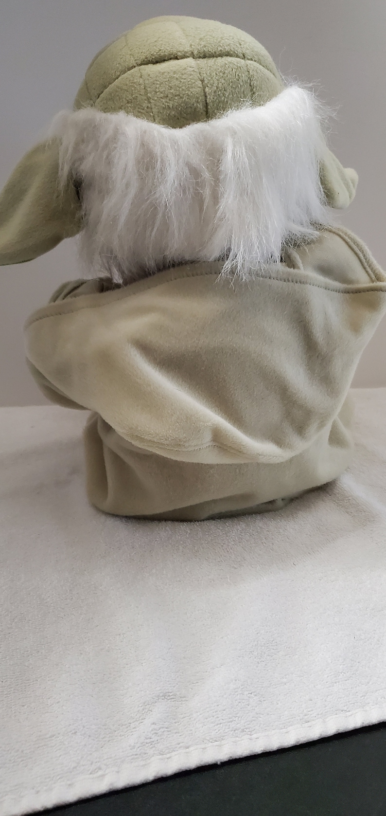 Op de afbeelding: Een pluche speelgoed van een groen personage met een witte bontkraag en een capuchon. Het speelgoed zit op een wit oppervlak.