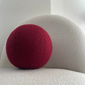 Pode incluir: Um grande travesseiro redondo vermelho com uma superfície texturizada está em um braço de cadeira branco texturizado.