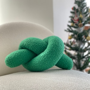 Puede incluir: Una almohada verde anudada se encuentra sobre una silla blanca y esponjosa. La almohada está hecha de un material suave y peludo.