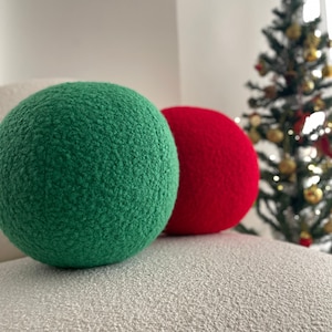 Op de afbeelding: Twee ronde, pluche kussens, een groen en een rood, liggen op een wit, getextureerd oppervlak. De kussens zijn zacht en pluche, en ze zouden perfect zijn om elke kamer een vleugje kleur en comfort te geven.