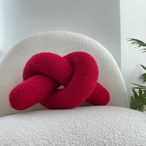 Puede incluir: Una almohada roja anudada, hecha de tela suave y peluda, se encuentra sobre una silla blanca texturizada.