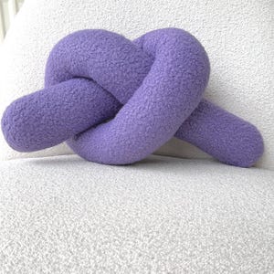 May include: A purple knot cushion made of a textured, fuzzy fabric. The cushion is shaped into a continuous, looped knot design with rounded ends, showcasing a soft, tactile surface that appears plush and consistent in its vibrant violet colour.