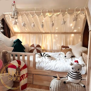 Op de afbeelding: Een houten bedframe met een witte kap en lichtslingers. Het bed is versierd met pluche speelgoed, waaronder een snoepstok, een vos en een gestreept dier. Het bed heeft een wit matras en een wit laken.