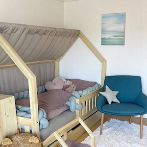 May include: A wooden framed children's bed with a canopy, featuring a pink duvet and a heart-shaped pillow. A blue armchair with a star cushion is next to the bed. A painting of the sea hangs on the wall. The room has a soft, inviting atmosphere.